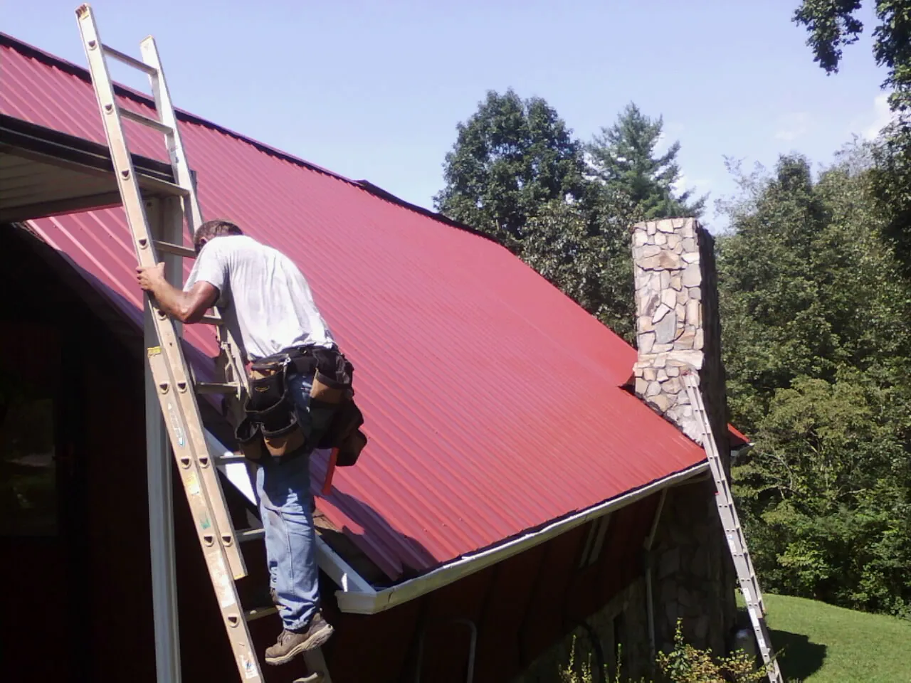 Roofer on a ladder beside a red metal roof and stone chimney — Soffit & Fascia Repair by Roofing Brothers in Galt